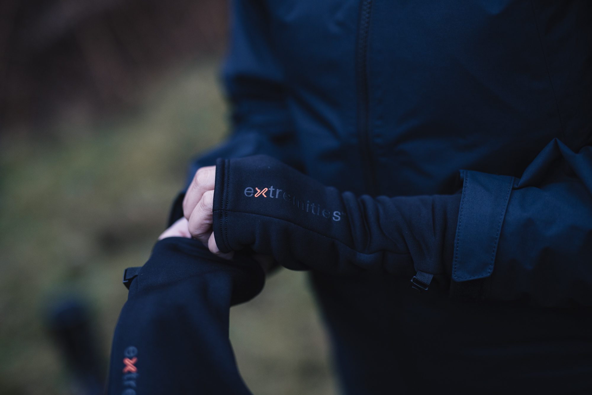 Close-up of two hands adjusting black thumb-hole gloves with visible "extremities" branding over a dark waterproof jacket against a blurred outdoor background.