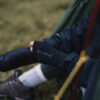 Power Liner Wrist Gaiter (2) Close-up of a person sitting on grass, wearing a dark jacket and gloves, with a focus on their hands and lower legs, showcasing outdoor gear and a relaxed camping atmosphere.