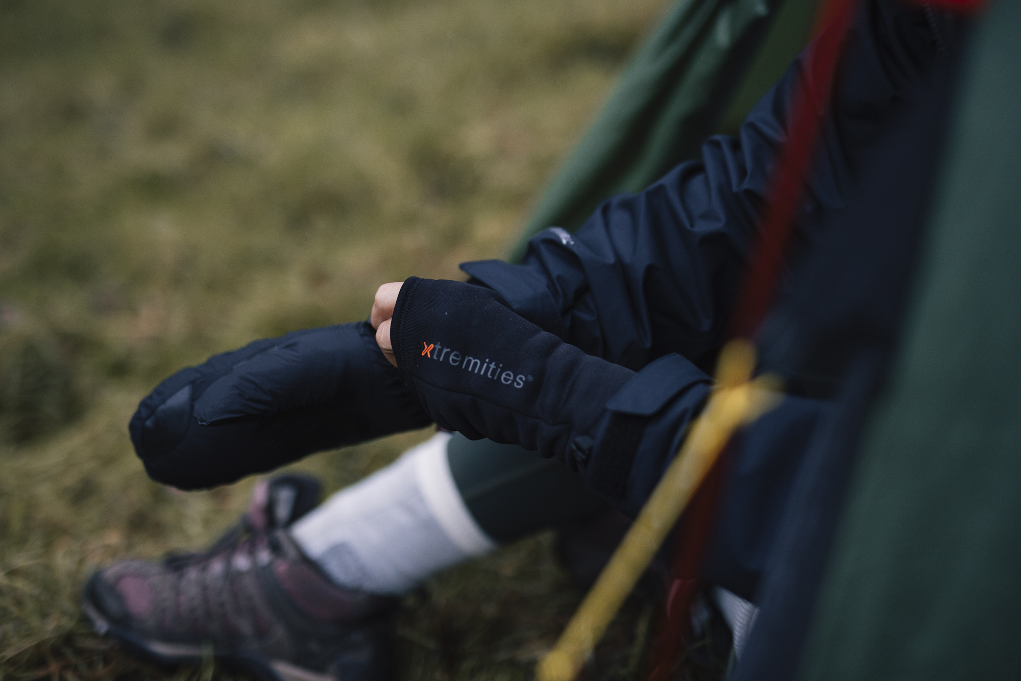 Close-up of a person sitting on grass, wearing a dark jacket and gloves, with a focus on their hands and lower legs, showcasing outdoor gear and a relaxed camping atmosphere.