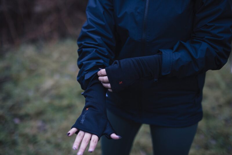 Person outdoors wearing a navy jacket and black thumb‑hole gloves, adjusting one sleeve with their hands visible against a blurred grassy background.