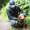 Quest-glove-with-dog-2500px-square-1.jpg A man in a dark jacket and cap crouching on a woodland path beside a reddish-brown spaniel with damp fur, holding the dog's blue lead as ferns fill the background.