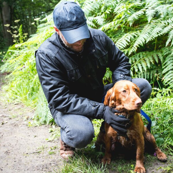 Quest-glove-with-dog-2500px-square-1.jpg A man in a dark jacket and cap crouching on a woodland path beside a reddish-brown spaniel with damp fur, holding the dog's blue lead as ferns fill the background.