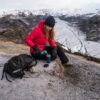SC102396-1.jpg Hiker in a red down jacket and black beanie sits on a frosty ridge above a braided glacial valley, pouring a hot drink from a teal thermos into a blue cup beside a black backpack.
