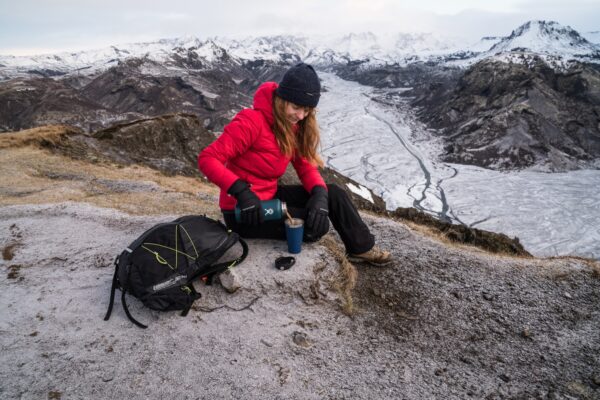 SC102396-1.jpg Hiker in a red down jacket and black beanie sits on a frosty ridge above a braided glacial valley, pouring a hot drink from a teal thermos into a blue cup beside a black backpack.
