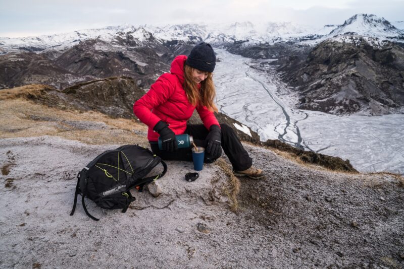 SC102396-1.jpg Hiker in a red down jacket and black beanie sits on a frosty ridge above a braided glacial valley, pouring a hot drink from a teal thermos into a blue cup beside a black backpack.