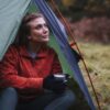 Silk-Liner-gloves-11lr-3.jpg Woman in a red zip-up jacket and black gloves sits at the opening of a green tent, holding a mug and gazing upward toward an autumn woodland clearing.