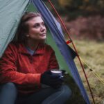 Woman in a red zip-up jacket and black gloves sits at the opening of a green tent, holding a mug and gazing upward toward an autumn woodland clearing.