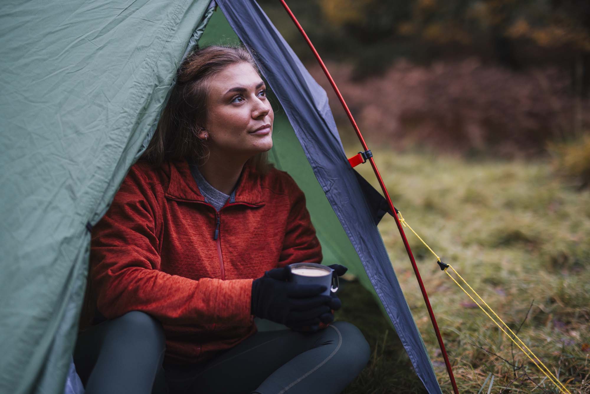 Woman in a red zip-up jacket and black gloves sits at the opening of a green tent, holding a mug and gazing upward toward an autumn woodland clearing.