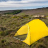 Terra Nova Shoot A bright yellow two-person tent pitched on a grassy moor dotted with white cotton‑grass, with a low rocky escarpment and layered cloudy sky on the horizon.