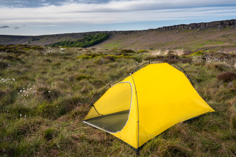 Terra Nova Shoot A bright yellow two-person tent pitched on a grassy moor dotted with white cotton‑grass, with a low rocky escarpment and layered cloudy sky on the horizon.