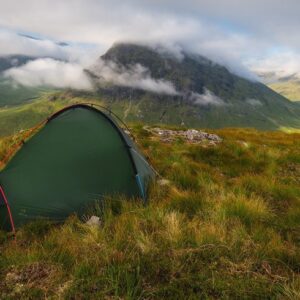 Green two-person tent pitched on a grassy, rocky ridge overlooking a valley with mist-shrouded mountains and low clouds in soft morning light