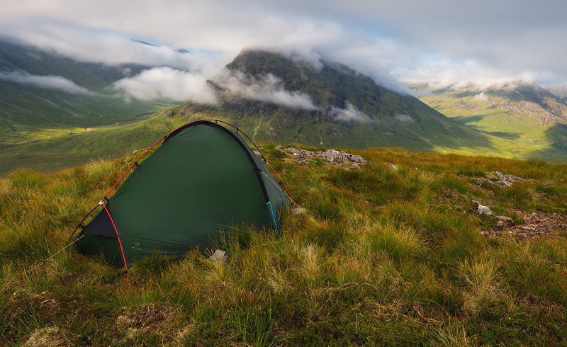 Green two-person tent pitched on a grassy, rocky ridge overlooking a valley with mist-shrouded mountains and low clouds in soft morning light