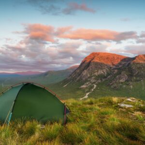 Green tent pitched on a grassy hillside overlooking a broad valley and rugged mountains bathed in warm sunset light beneath pink-tinged clouds.