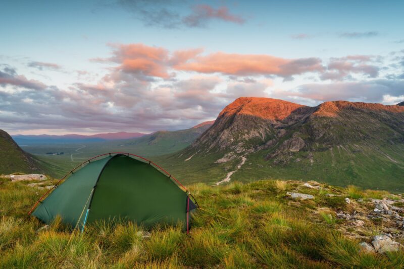 Green tent pitched on a grassy hillside overlooking a broad valley and rugged mountains bathed in warm sunset light beneath pink-tinged clouds.