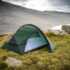 Southern-Cross-2-2023-LR-3.jpg A green dome tent with red poles and yellow guylines is pitched on a wind‑swept grassy slope beside lichen‑covered rocks, its door open to reveal a pale inner sleeping area, with a lake and misty hills in the background under a cloudy sky.