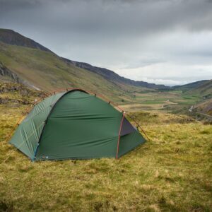 Green dome tent pitched on a grassy hillside with guy lines staked down, overlooking a broad valley and winding road between rugged mountains beneath a cloudy, overcast sky.