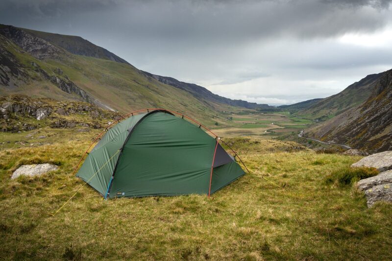 Southern-Cross-2-LR-3.jpg Green dome tent pitched on a grassy hillside with guy lines staked down, overlooking a broad valley and winding road between rugged mountains beneath a cloudy, overcast sky.