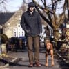 Sportsman-and-Woburn-72dpi-3.jpg Man walking a brown short-coated dog on a lead along a village pavement, dressed in a dark quilted jacket, flat cap, gloves, brown trousers and leather boots, with stone buildings and bare trees in the sunlit background.