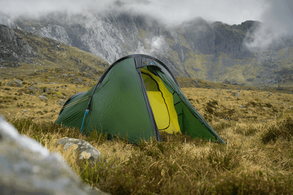 Green dome tent with a partially open door showing a bright yellow inner layer, pegged with guylines on windswept grassy moorland beneath mist-shrouded rocky mountains.