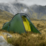 Green dome tent with a partially open door showing a bright yellow inner layer, pegged with guylines on windswept grassy moorland beneath mist-shrouded rocky mountains.