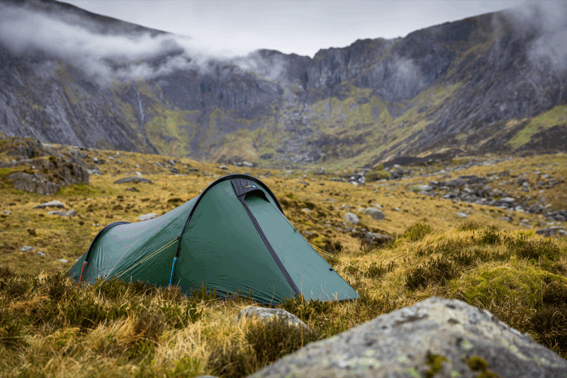 Starlite-1-1_LR-1.png A green backpacking tent pitched on grassy, rocky moorland with steep, mist-shrouded mountains and low grey clouds in the background.