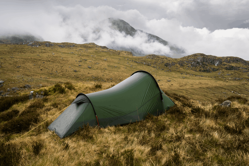 Starlite-1-23_LR-1.png A lone olive-green tunnel tent pitched on windswept brown grass amid rocky moorland, with low cloud and mist rolling over distant mountains under an overcast sky.