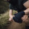 Station-Gloves-new4-3.jpg Crouching person in dark athletic clothing and a neck gaiter with gloved hands clasped over a knee on a dirt trail, showing muscular forearms and a blurred grassy background.