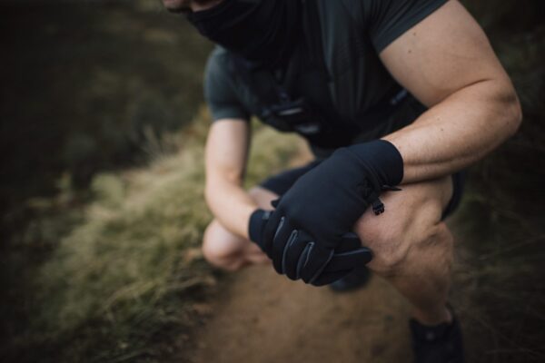 Station-Gloves-new4-3.jpg Crouching person in dark athletic clothing and a neck gaiter with gloved hands clasped over a knee on a dirt trail, showing muscular forearms and a blurred grassy background.