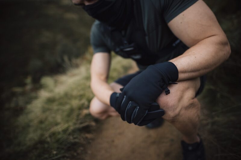 Station-Gloves-new4-3.jpg Crouching person in dark athletic clothing and a neck gaiter with gloved hands clasped over a knee on a dirt trail, showing muscular forearms and a blurred grassy background.