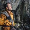 Station-gloves-6-LR-3.jpg Man in an orange jacket and gloves stands inside a rocky cave holding coiled climbing rope over his shoulder, wearing a climbing harness and looking up at the cliff face.
