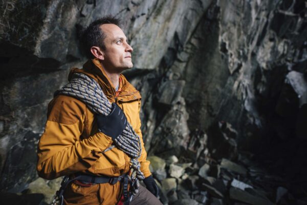 Station-gloves-6-LR-3.jpg Man in an orange jacket and gloves stands inside a rocky cave holding coiled climbing rope over his shoulder, wearing a climbing harness and looking up at the cliff face.