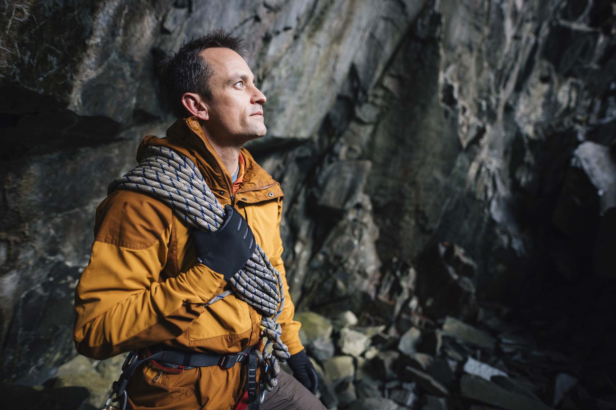 Station-gloves-6-LR-3.jpg Man in an orange jacket and gloves stands inside a rocky cave holding coiled climbing rope over his shoulder, wearing a climbing harness and looking up at the cliff face.