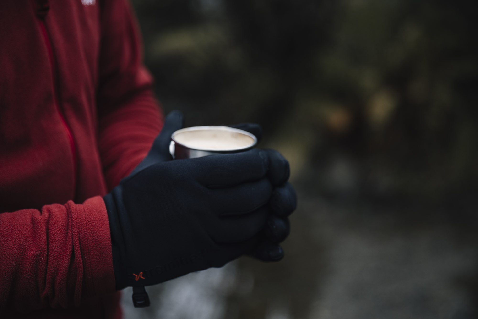 Person in a red fleece jacket wearing black gloves holding a small metal cup of creamy hot drink against a blurred outdoor background