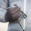 Close-up of a hand in a black winter glove gripping the handle and strap of a trekking pole against a grey padded jacket background.