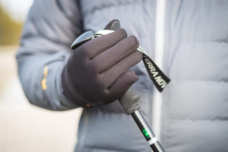 Sticky-Waterproof-Power-Liner-Glove-5-scaled-2.jpg Close-up of a hand in a black winter glove gripping the handle and strap of a trekking pole against a grey padded jacket background.