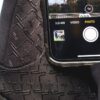 Close-up of a smartphone resting on textured black fabric with a raised plus-shaped pattern, showing the camera app in Photo mode with a white circular shutter, 1x zoom indicator, a small landscape preview and a faint crack across the phone's glass.