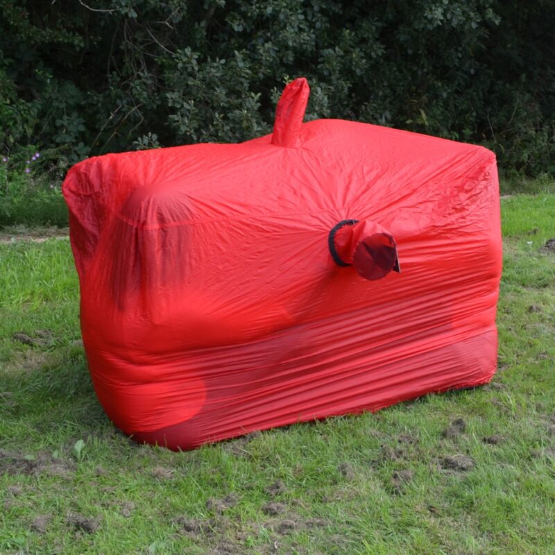 Superlight-Bothy-2-in-use-square-1500px-1.jpg Bright red stretch fabric tightly wrapped around a large rectangular object on grass, with a small round nozzle-like protrusion on the front and a short vertical bulge on top, set against a backdrop of trees.