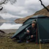 Woman sitting at the entrance of a dark green Pioneer Lite 2 tent by a calm mountain lake, pouring a hot drink from a Jetboil stove into a mug beneath overcast skies.
