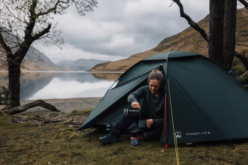 Woman sitting at the entrance of a dark green Pioneer Lite 2 tent by a calm mountain lake, pouring a hot drink from a Jetboil stove into a mug beneath overcast skies.