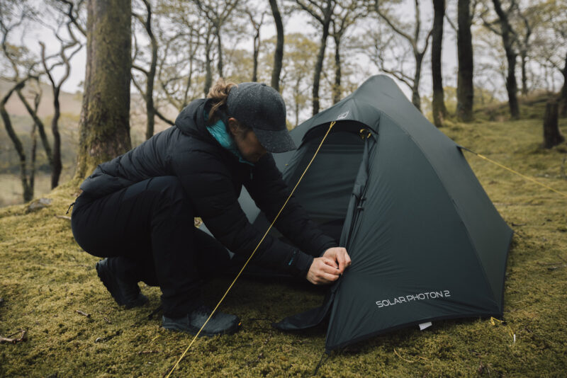 TERRA_NOVA_PITCHING_APRIL25_0056 A person in outdoor gear is setting up a green camping tent in a forested area, demonstrating proper tent assembly techniques in a natural setting.