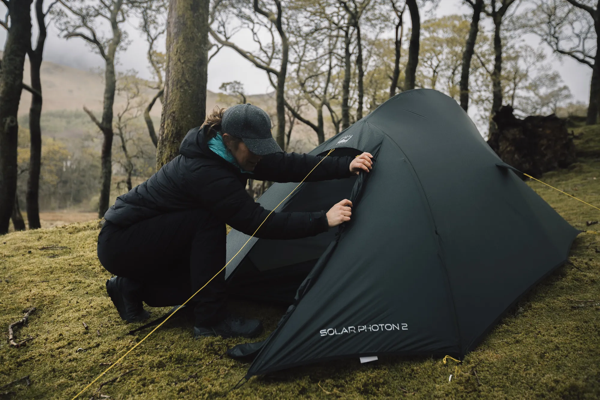 Person setting up a green camping tent in a forested area, surrounded by trees and grass, showcasing outdoor adventure and camping skills.