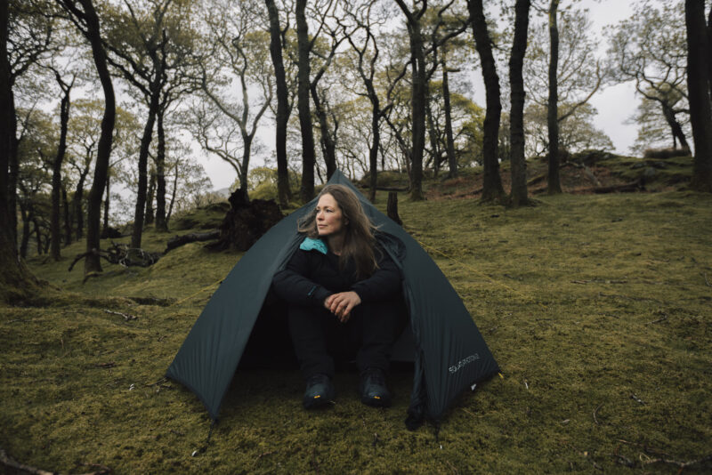 TERRA_NOVA_PITCHING_APRIL25_0059 Woman sitting in front of a tent in a forested area, surrounded by trees, showcasing outdoor camping and adventure.