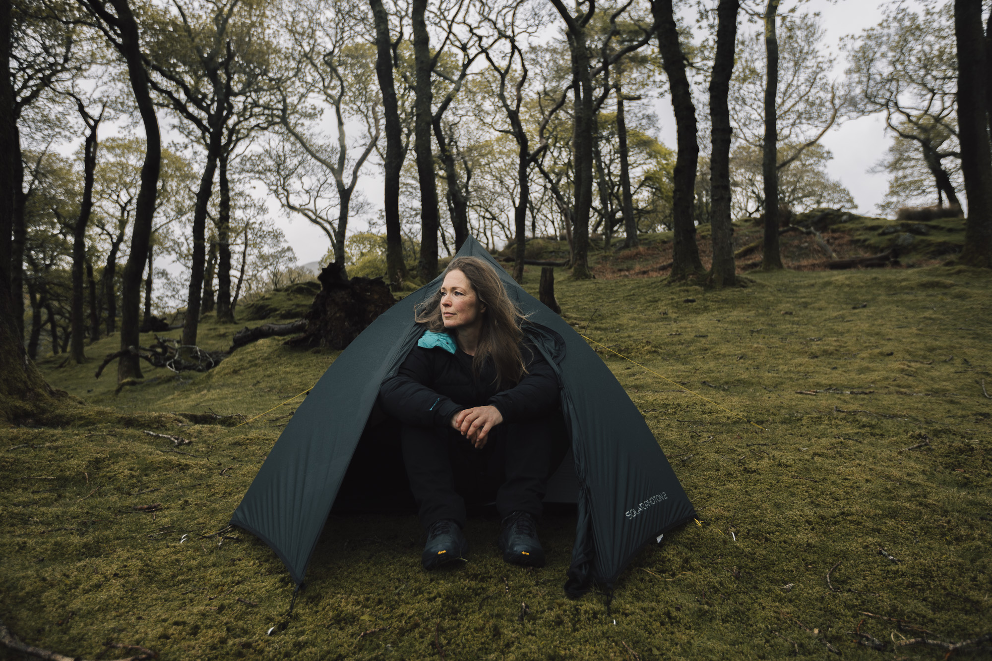 Woman sitting in front of a tent in a forested area, surrounded by trees, showcasing outdoor camping and adventure.