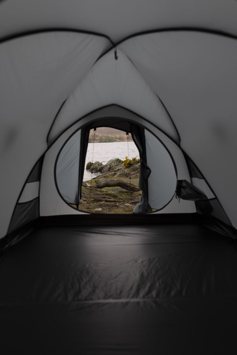 View from inside a grey tent looking out through the open entrance onto a rocky lakeshore with a fallen log, yellow gorse flowers at the water's edge and hills across the calm water.