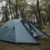 Teal dome tent with its front flaps partially open, pitched on damp grass in a misty deciduous woodland surrounded by tall, leafed trees.