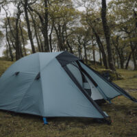 Teal dome tent with its front flaps partially open, pitched on damp grass in a misty deciduous woodland surrounded by tall, leafed trees.