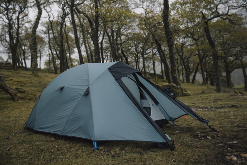 Teal dome tent with its front flaps partially open, pitched on damp grass in a misty deciduous woodland surrounded by tall, leafed trees.