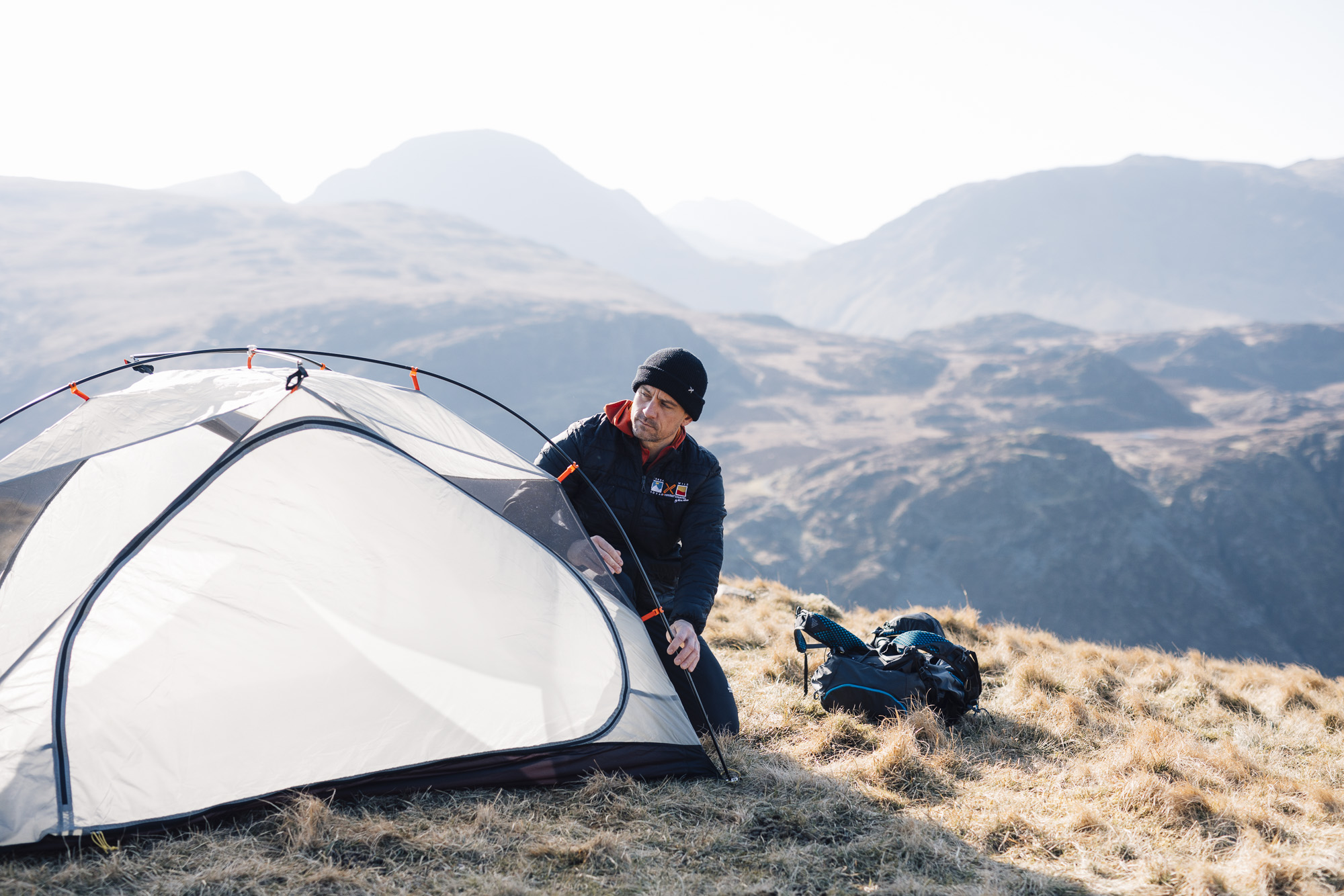 Person in a yellow-and-black jacket pitching a small white tent on a grassy ridge, with a backpack and gear nearby and rolling green hills under a pale sky in the background.