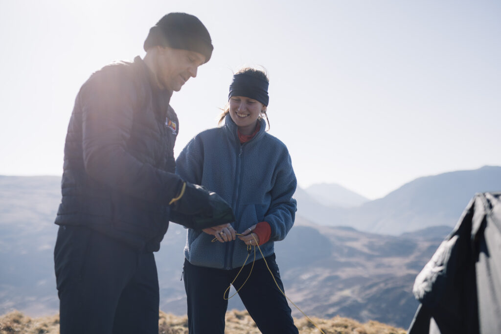 Two climbers preparing equipment outdoors in a mountainous landscape, with sunlight illuminating their faces and surrounding scenery.