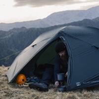 Bearded man in a black beanie crouches at the open door of a dark two-person tent on a grassy mountain ridge, preparing a hot drink on a small camping stove beside an orange climbing helmet and ropes, with layered rocky peaks in the misty background.